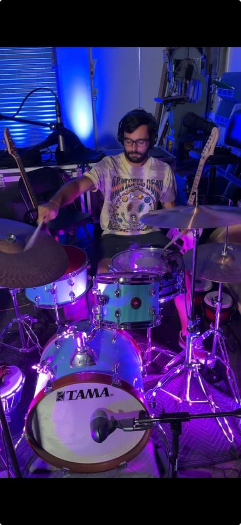 Mario Fantini playing a TAMA drum kit in the studio with blue and purple lighting.